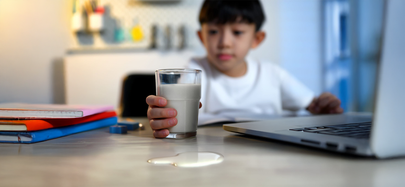 Header image: A student holding a glass of milk with a spill near a school laptop — highlighting the need for device protection.